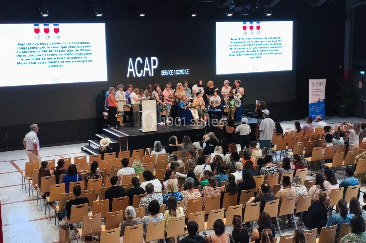 Un groupe de personnes sur scène devant un public assis, avec des écrans affichant des informations en arrière-plan.