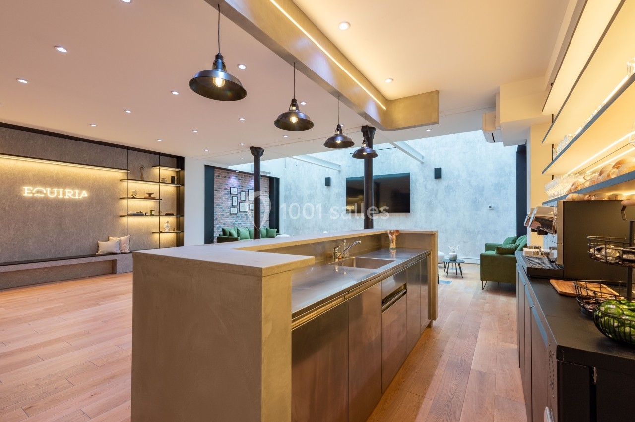 Cuisine moderne avec îlot central en béton, éclairage suspendu, parquet en bois clair et espace salon en arrière-plan.