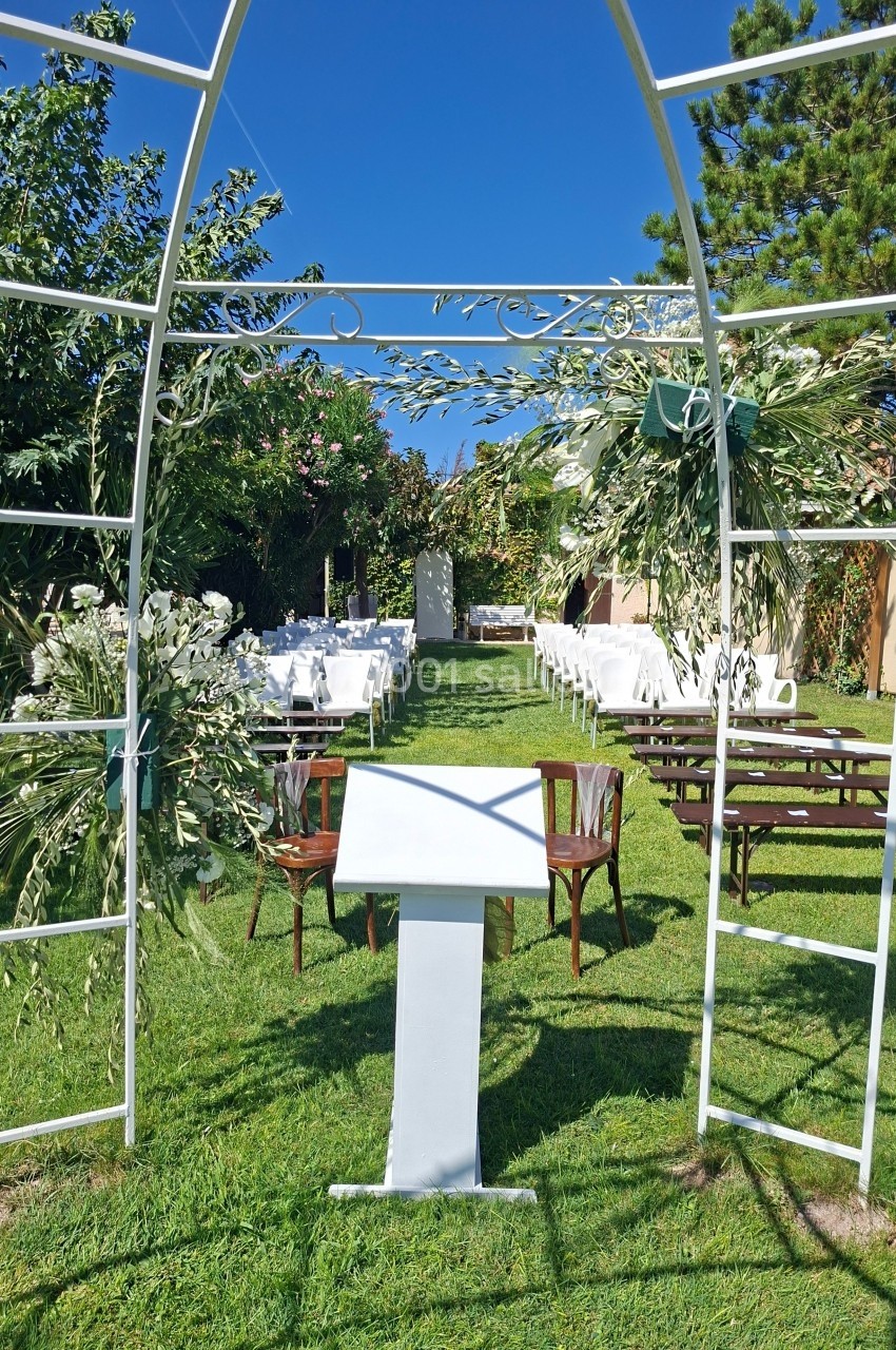 Arche blanche décorée de feuillages, avec des chaises alignées sur une pelouse pour une cérémonie en extérieur.