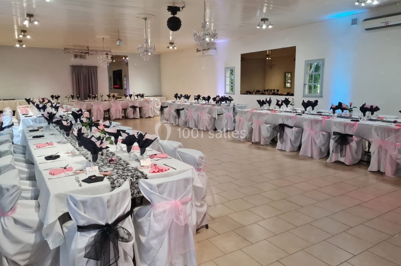 Salle de réception décorée avec des tables alignées, nappes blanches et nœuds noirs et roses sur les chaises.