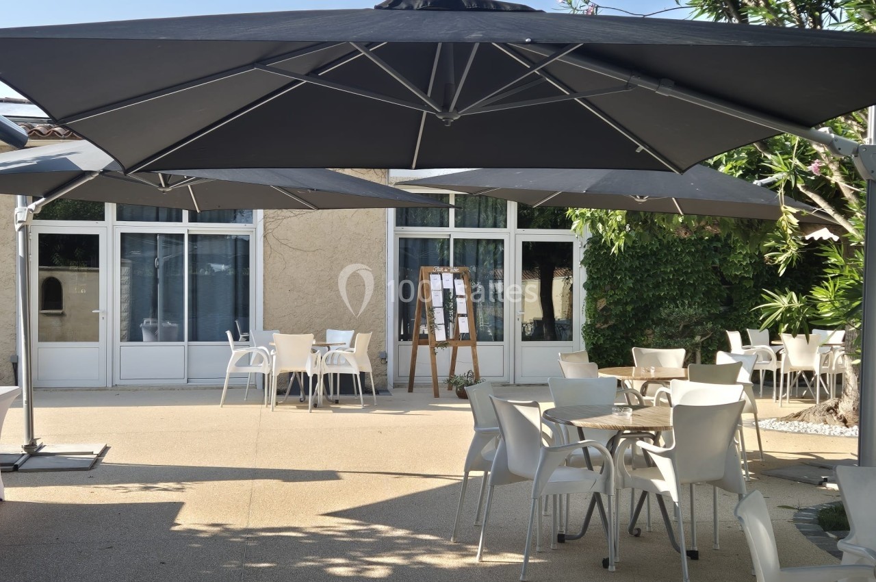 Terrasse extérieure avec tables et chaises blanches sous de grands parasols noirs, entourée de verdure.