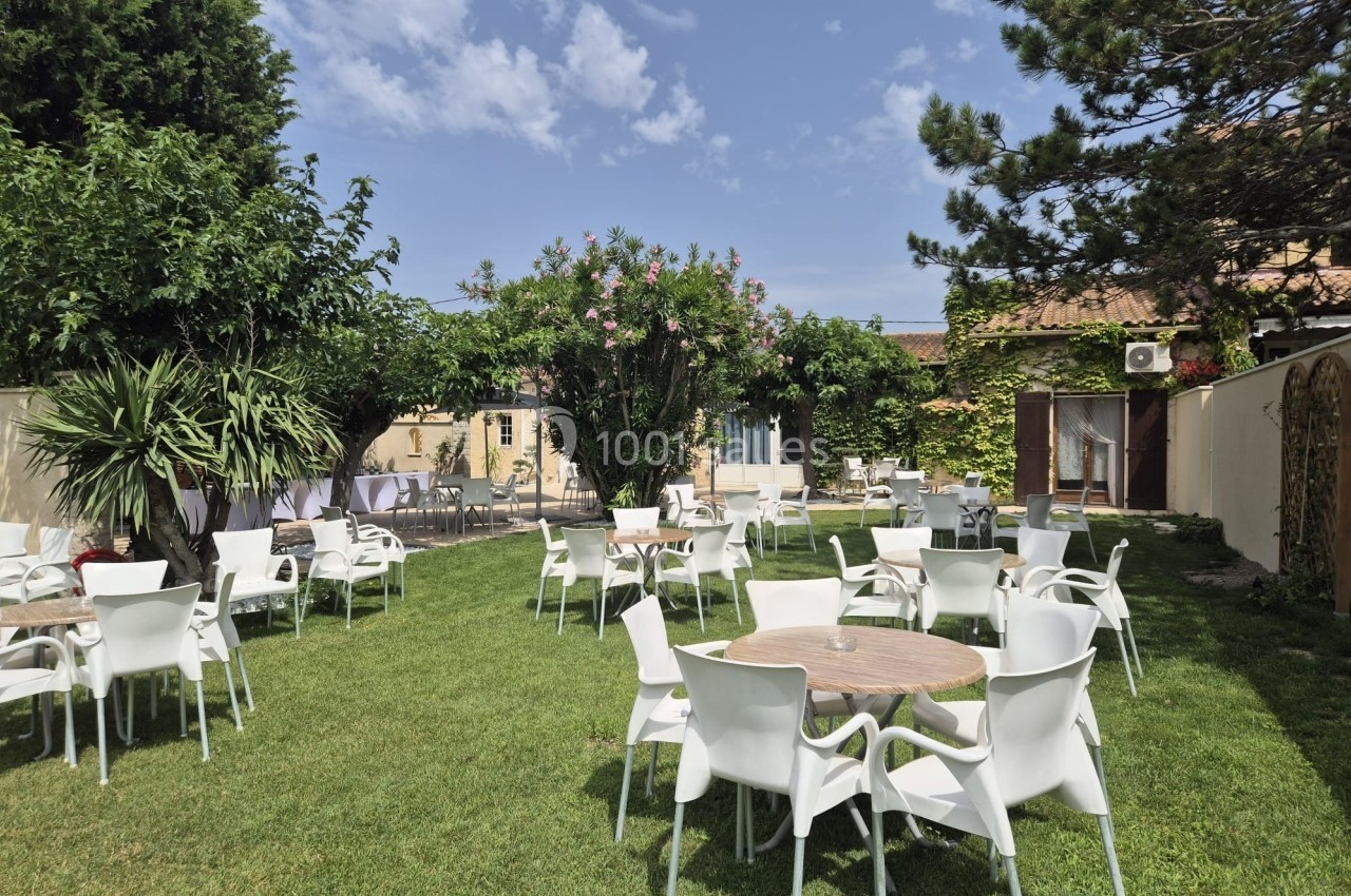 Jardin avec des tables et chaises blanches disposées sur une pelouse, entouré de végétation et bâtiments en arrière-plan.
