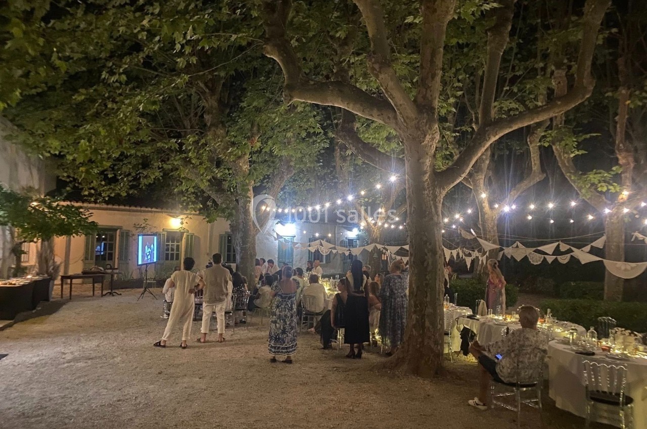 Groupe de personnes rassemblées en soirée dans un jardin éclairé par des guirlandes lumineuses, avec tables décorées.