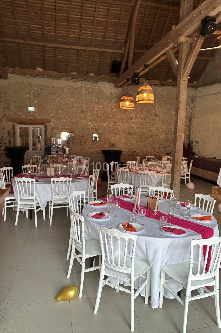 Salle de réception rustique avec tables rondes dressées, nappes blanches, chaises en bois et décorations simples.