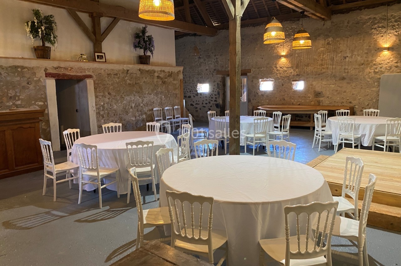 Salle rustique avec murs en pierre, tables rondes couvertes de nappes blanches et chaises blanches, éclairée par des…