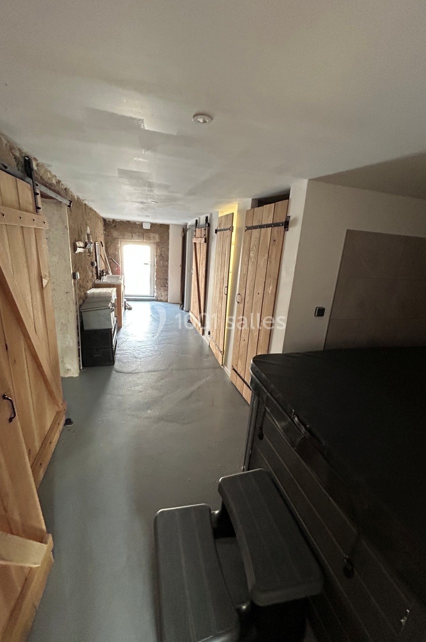 Couloir étroit avec sol en béton, murs en bois et pierre, portes coulissantes, et un jacuzzi noir au premier plan.