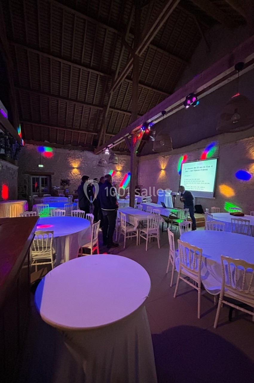 Salle de réception avec tables rondes et chaises blanches, éclairée par des lumières colorées et un projecteur.