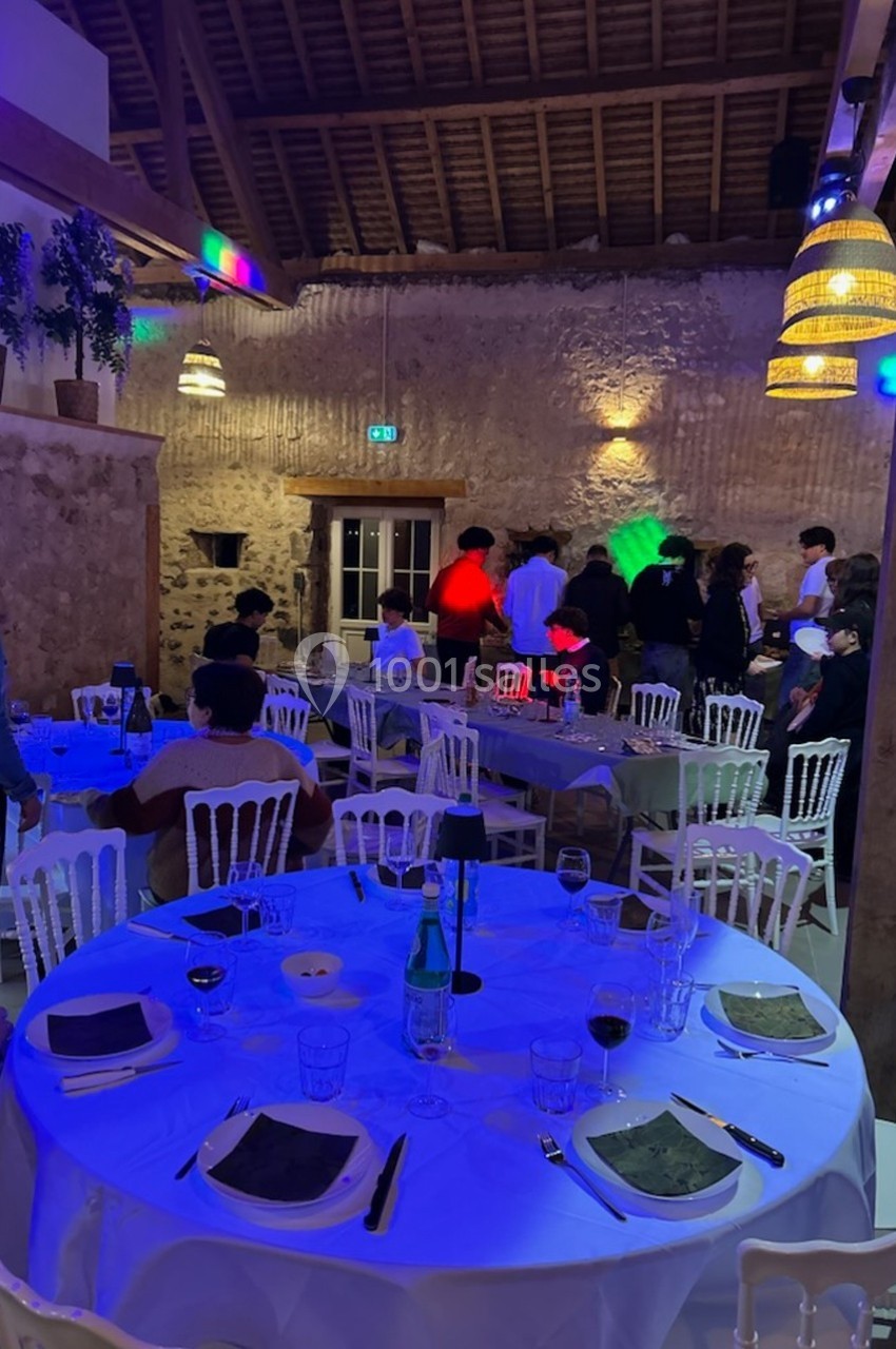 Salle de réception avec tables dressées, invités debout près d'un mur en pierre sous un éclairage coloré.