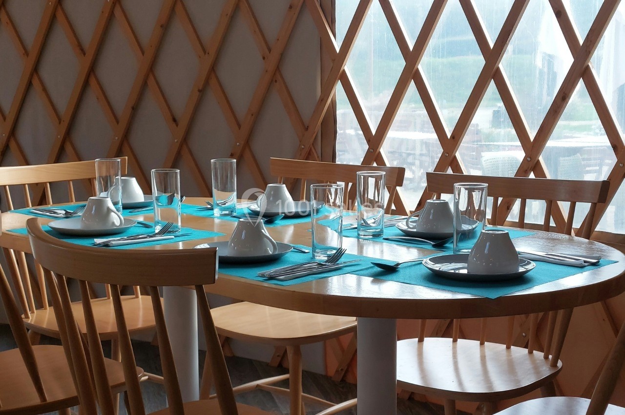 Table dressée avec des sets turquoise, verres, assiettes et couverts, entourée de chaises en bois dans une pièce lumineuse.