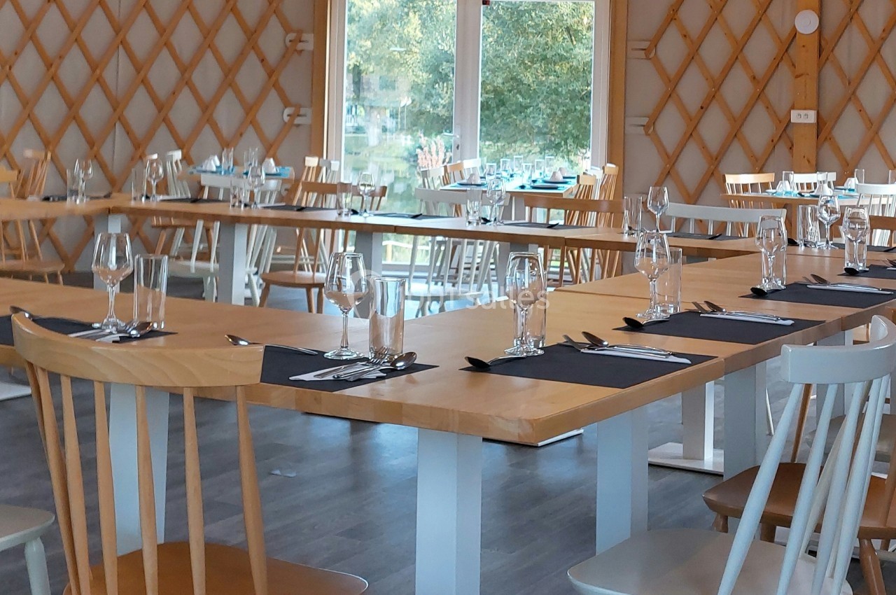 Salle de restaurant lumineuse avec tables en bois dressées, chaises variées et vue sur un extérieur verdoyant.