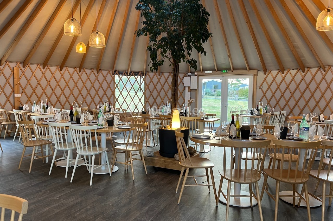 Salle circulaire en bois avec tables dressées, chaises dépareillées et un arbre central sous un toit en forme de yourte.
