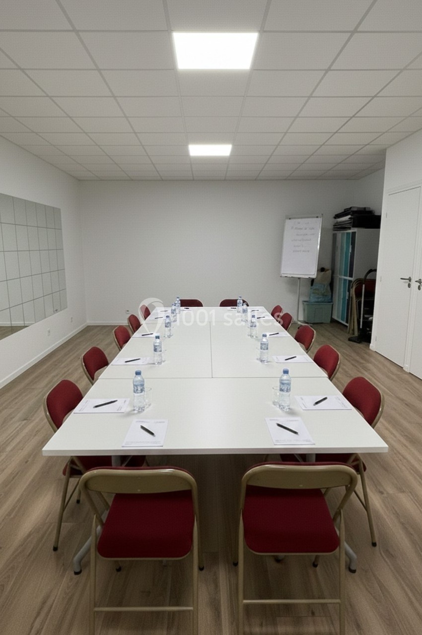 Salle de réunion avec une grande table rectangulaire, chaises rouges, bouteilles d'eau et carnets disposés pour chaque place.