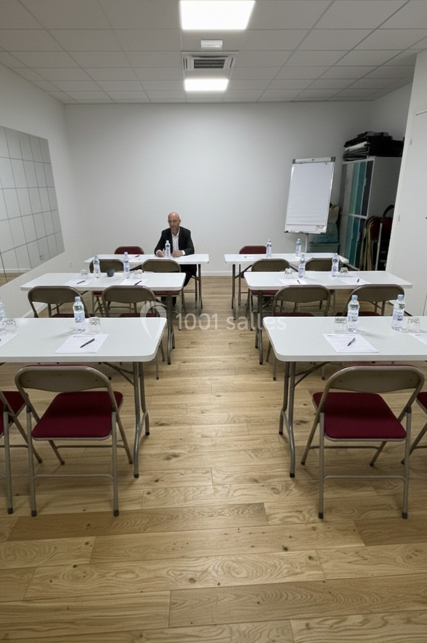Salle de réunion avec des tables et chaises alignées, un homme assis seul au fond, tableau blanc et miroir au mur.