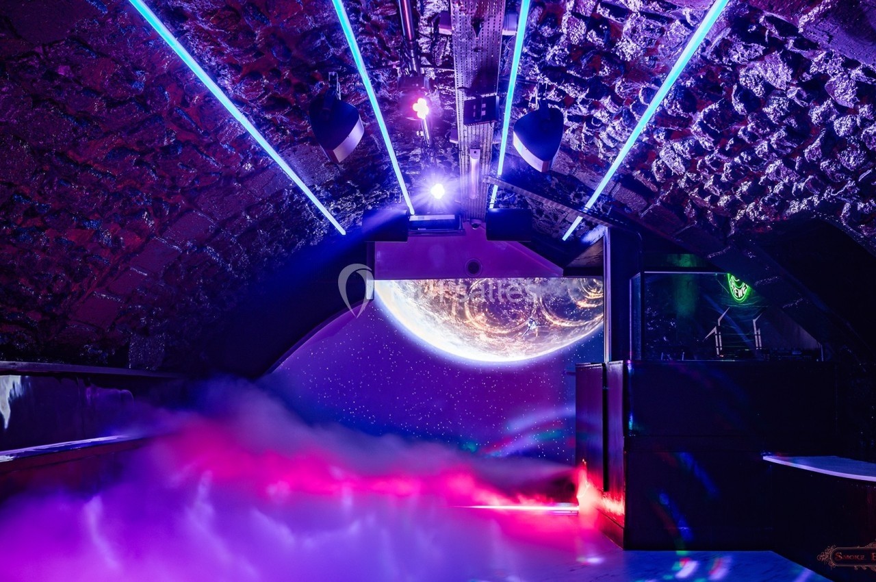 Salle voûtée avec éclairage néon coloré, fumée au sol et projection d'une planète sur un mur au fond.