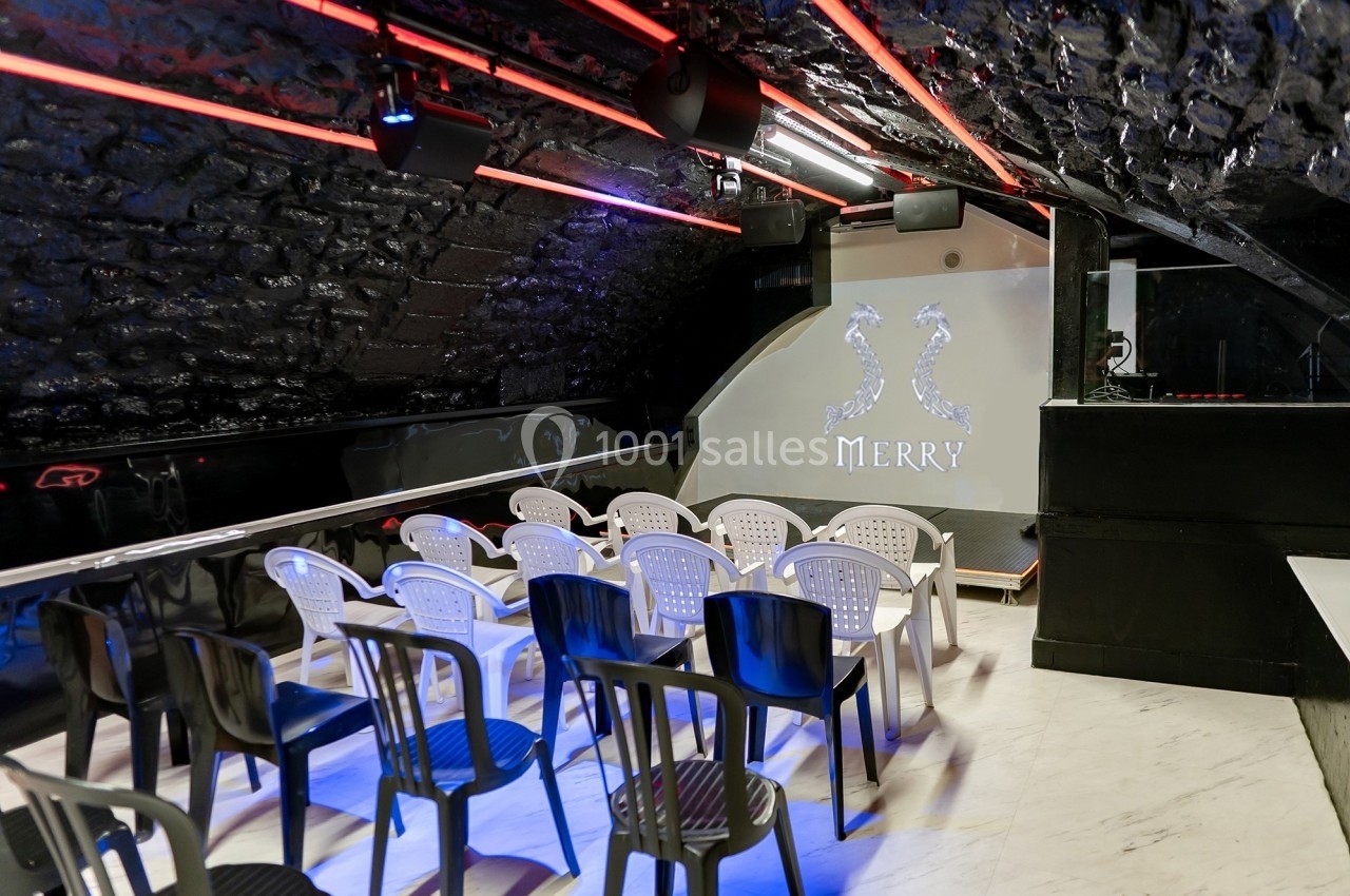Salle voûtée avec des chaises en plastique disposées face à une scène éclairée, ornée du logo ’Merry’.