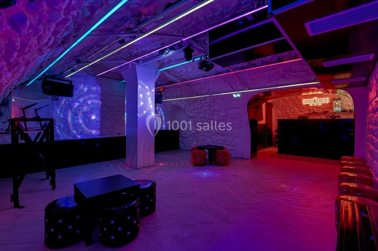 Salle voûtée avec murs en pierre, éclairage néon coloré, tabourets noirs et espace central dégagé.