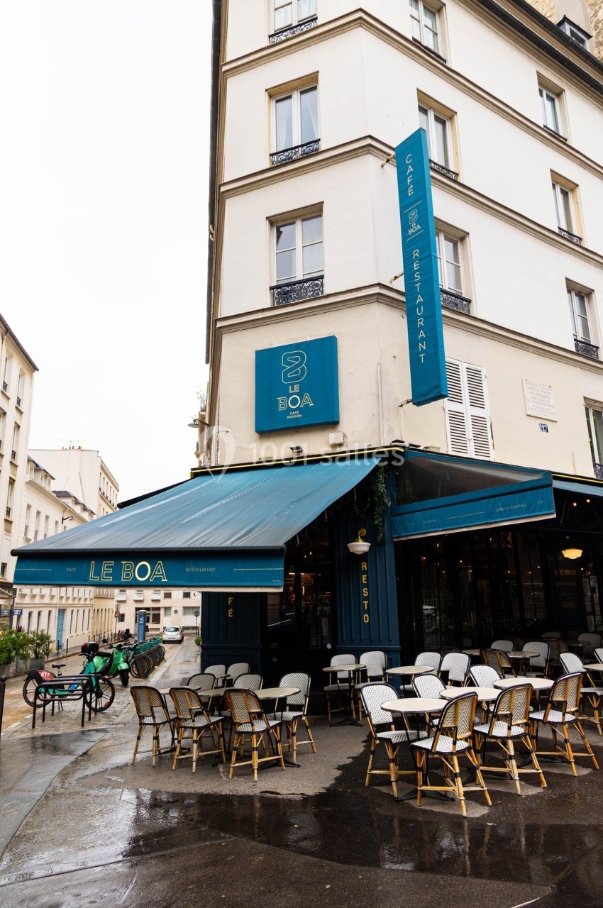 Façade d'un café-restaurant parisien avec terrasse extérieure, tables et chaises sous un auvent bleu.