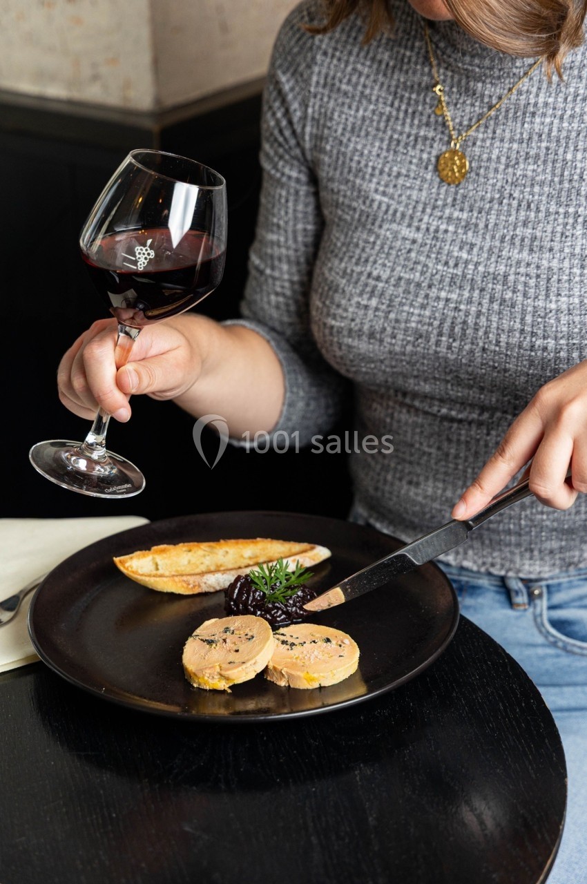 Une personne déguste du foie gras avec du pain grillé, accompagnée d'un verre de vin rouge sur une table sombre.