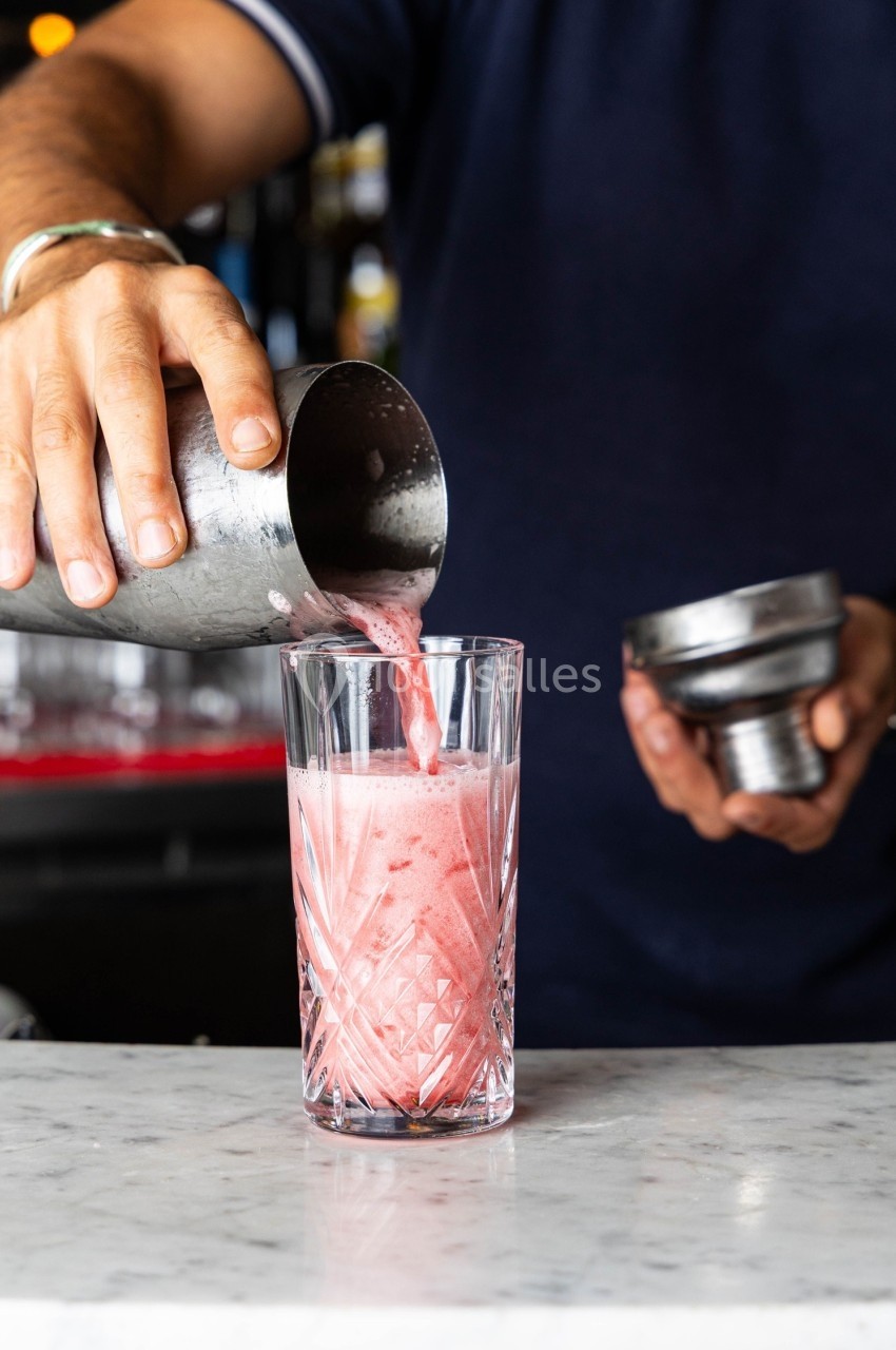 Un barman verse un cocktail rose mousseux d'un shaker dans un verre en cristal sur un comptoir en marbre.