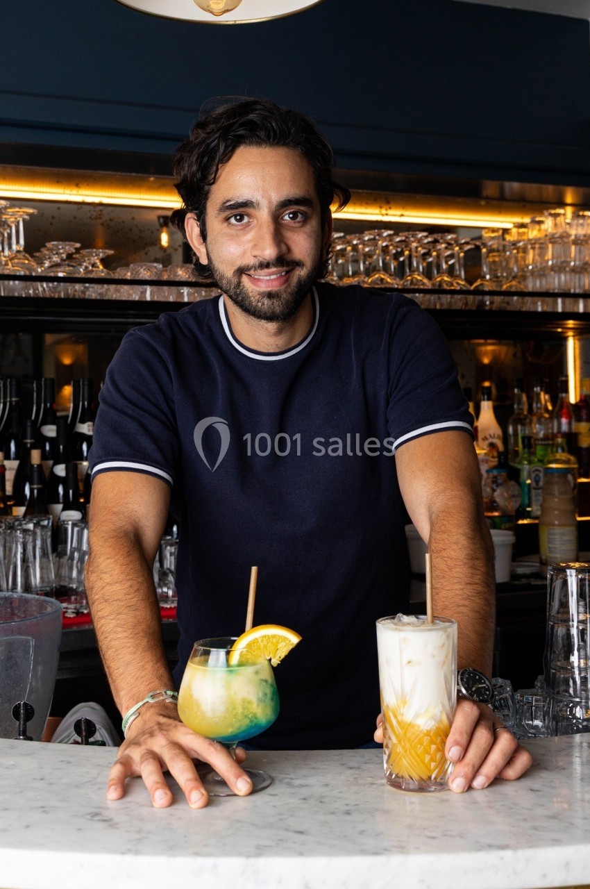 Un homme souriant derrière un comptoir de bar présente deux cocktails colorés sur une surface en marbre.