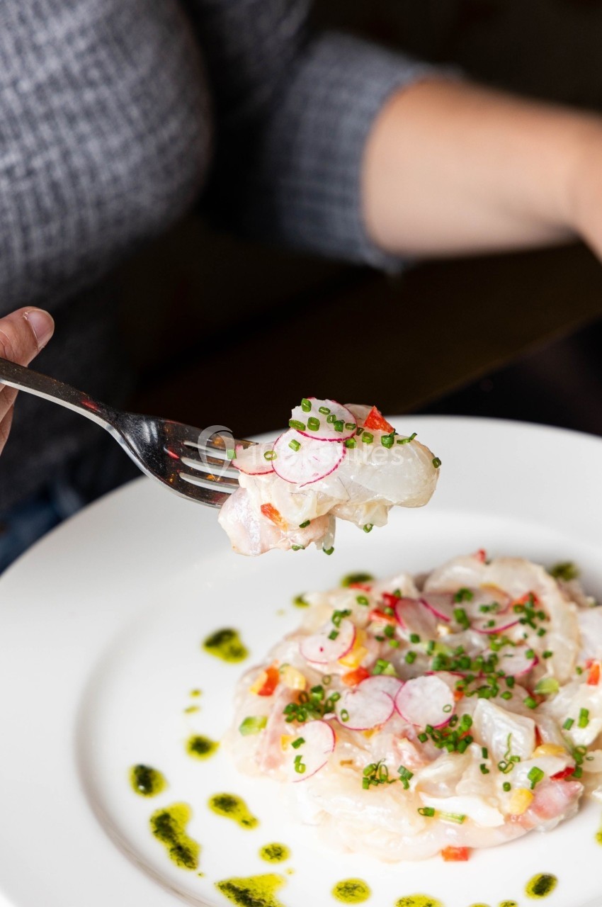 Assiette de carpaccio de poisson garnie de radis, ciboulette et huile verte, avec une fourchette en gros plan.