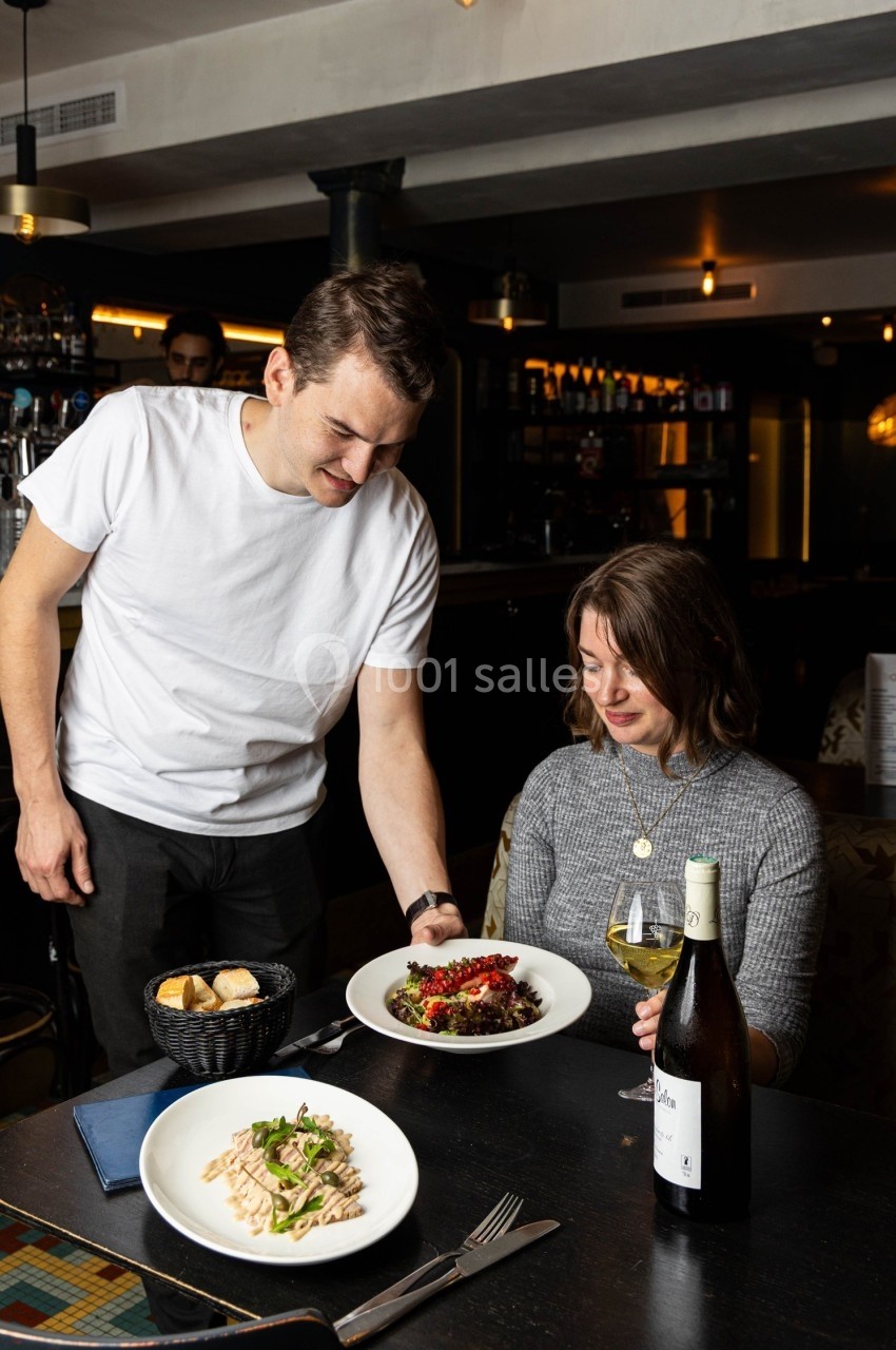 Un serveur sert un plat à une cliente assise dans un restaurant, avec des assiettes et une bouteille de vin sur la table.