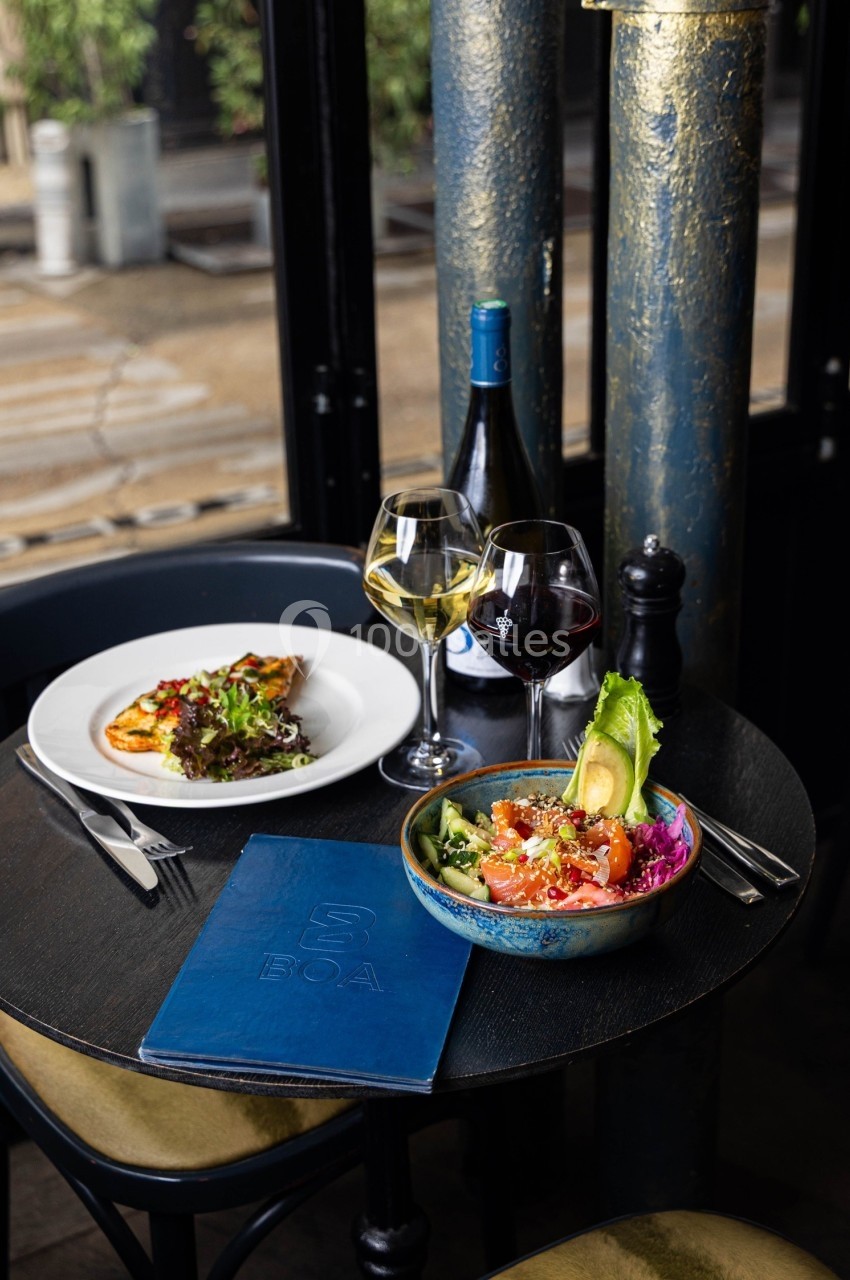 Table dressée avec un bol de poke coloré, une assiette de poisson, deux verres de vin et une bouteille en arrière-plan.