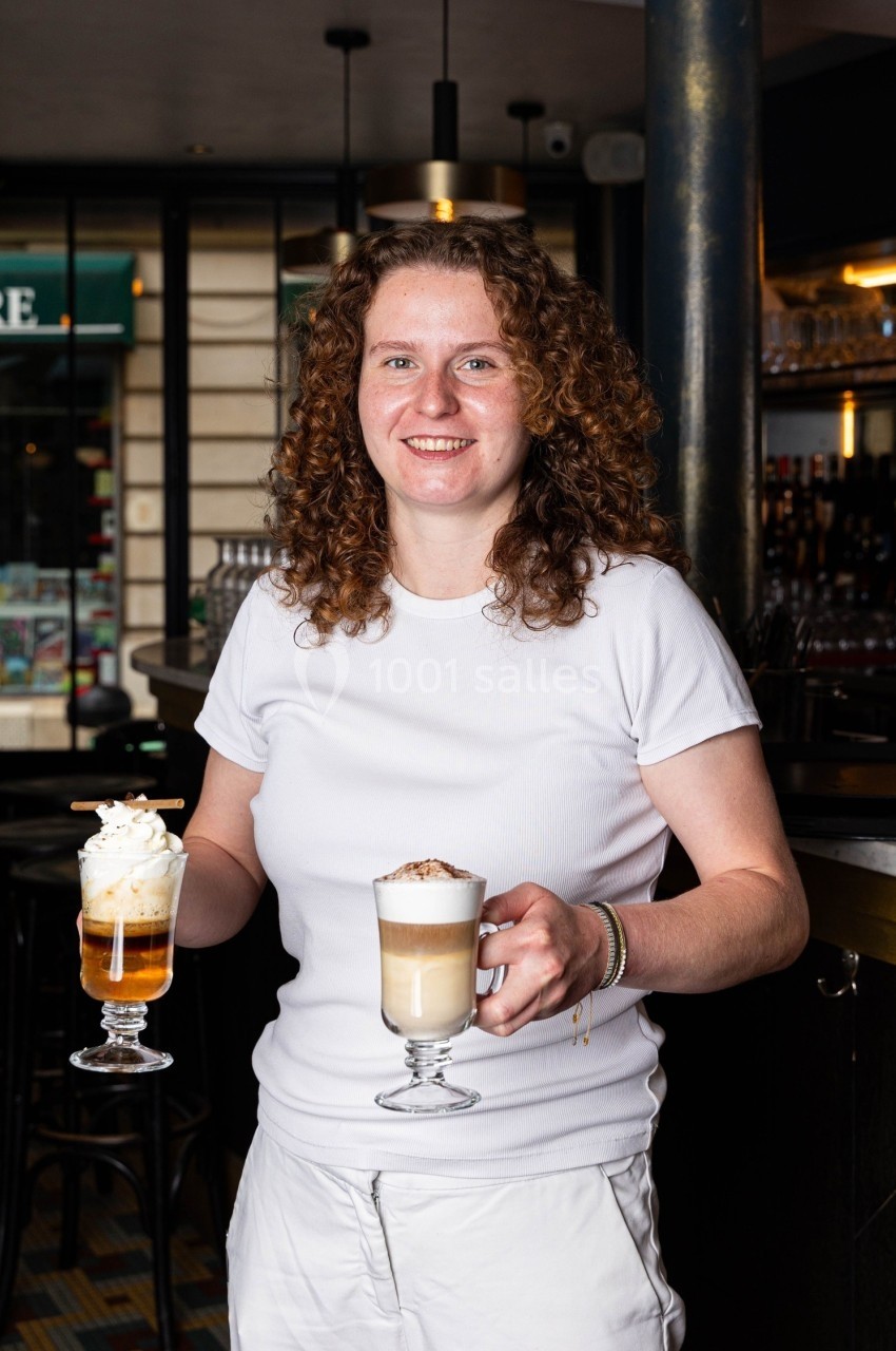 Une femme souriante tient un café latte et une boisson avec chantilly dans un café au décor chaleureux.