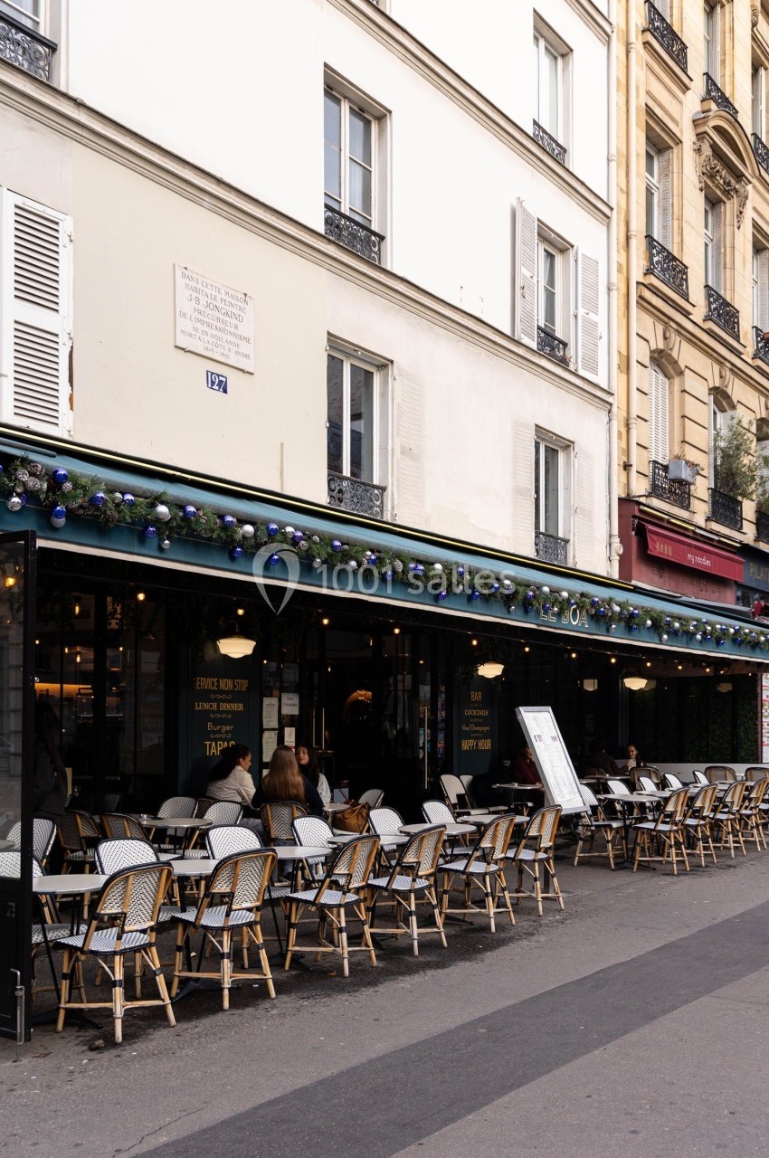 Terrasse d'un café parisien avec des chaises en osier, située au rez-de-chaussée d'un immeuble blanc.