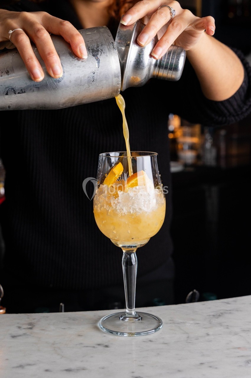 Une personne verse un cocktail jaune avec des glaçons et des zestes d'orange dans un verre à pied.