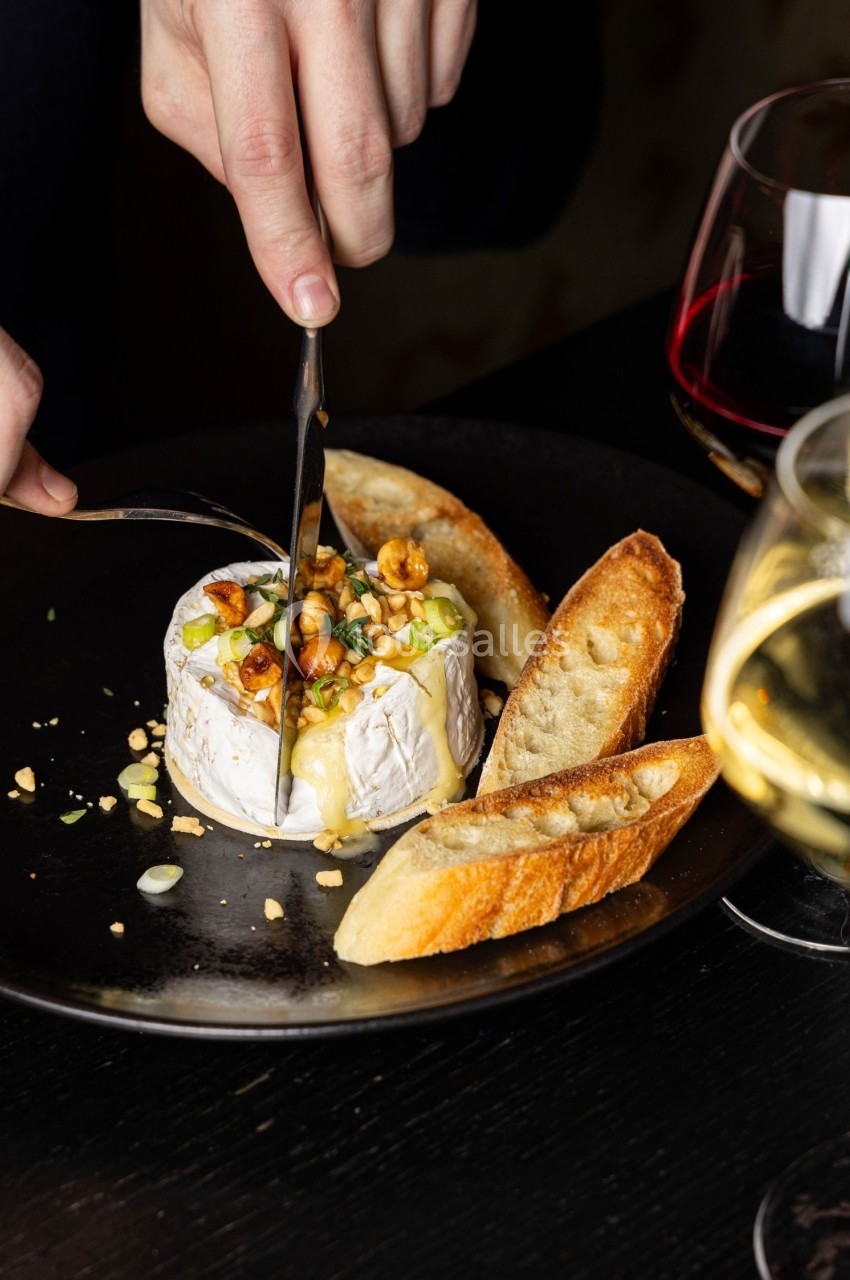 Une personne coupe un fromage fondu garni de noix et herbes, accompagné de tranches de pain et de verres de vin.