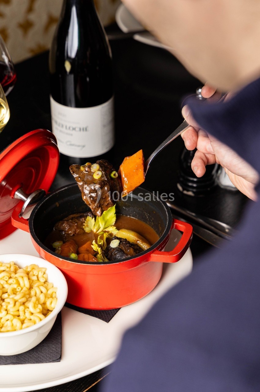 Une personne déguste un plat mijoté servi dans une cocotte rouge, accompagné de pâtes et de vin.