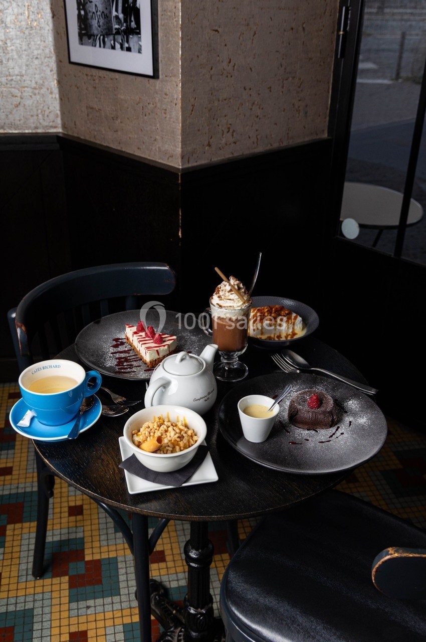 Table ronde avec desserts variés, théière, tasse de café et chocolat chaud dans un café au sol carrelé.