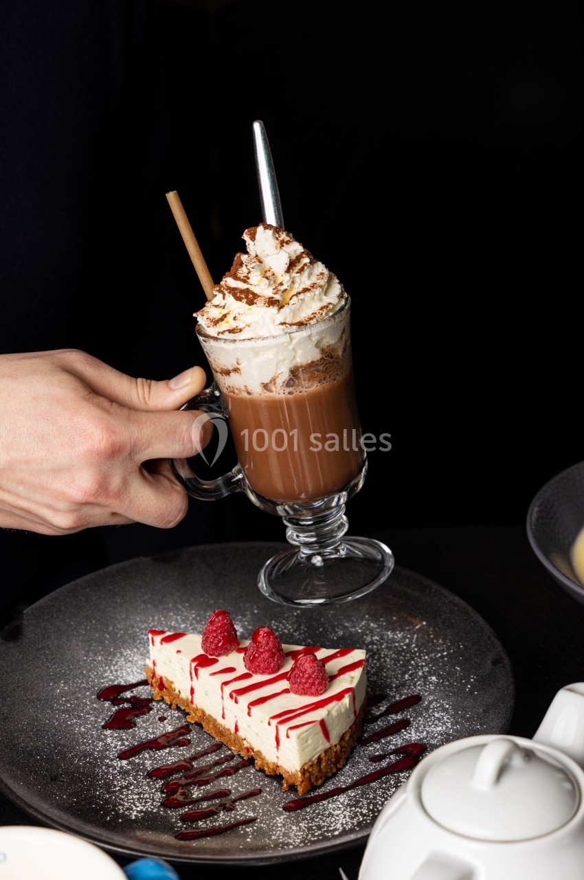 Une main tient un verre de chocolat chaud avec chantilly, posé à côté d'une part de gâteau décorée de framboises.