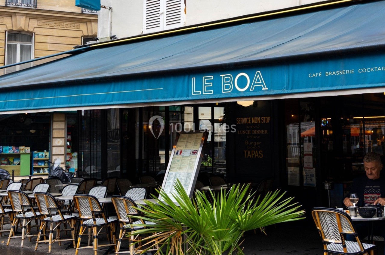 Terrasse d'un café-restaurant parisien avec des tables en osier, une devanture bleue et un menu affiché à l'entrée.