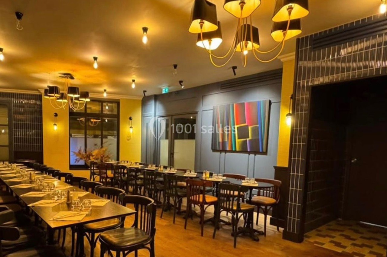 Salle de restaurant chaleureuse avec tables en bois alignées, chaises noires, éclairage tamisé et tableau coloré au mur.