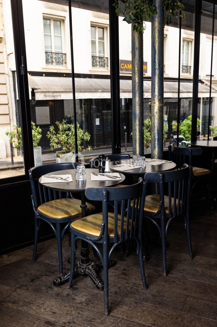 Tables rondes avec chaises en bois noir dans un café lumineux, donnant sur une rue bordée de vitrines et de plantes.