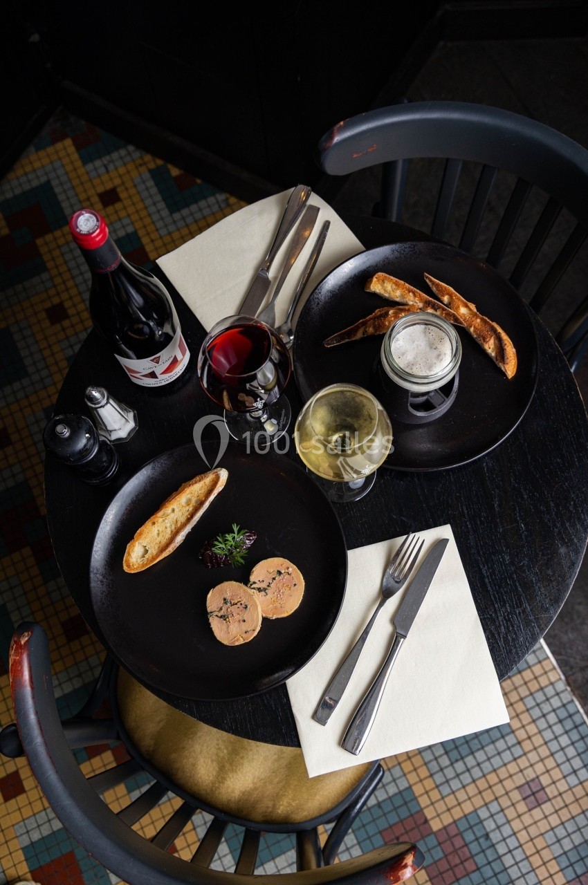 Table ronde avec assiettes de foie gras, pain grillé, verres de vin et bière, sur un sol en mosaïque colorée.