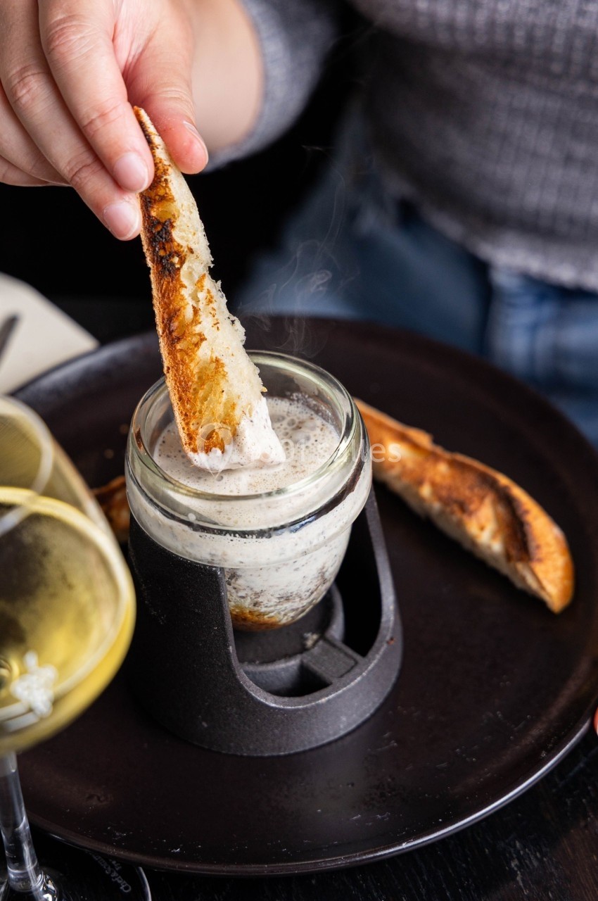 Une main trempe un morceau de pain grillé dans un pot contenant une préparation crémeuse, sur une table de restaurant.