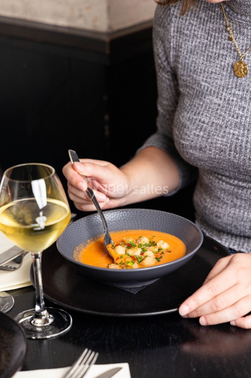 Une personne déguste une soupe orange garnie de croûtons et d'herbes, avec un verre de vin blanc à côté.