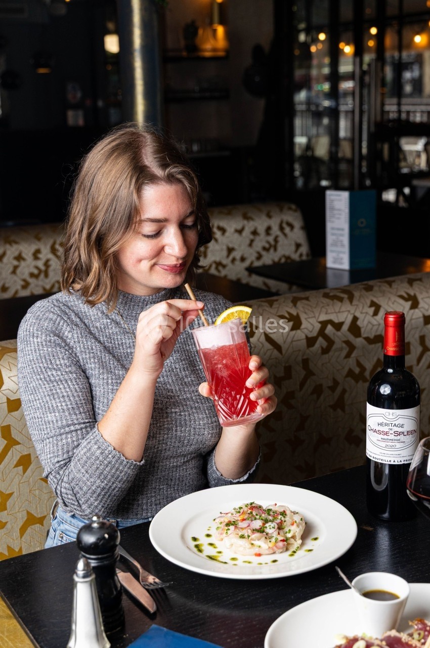 Une femme boit un cocktail rose avec une rondelle d'orange, assise à une table avec un plat et une bouteille de vin.