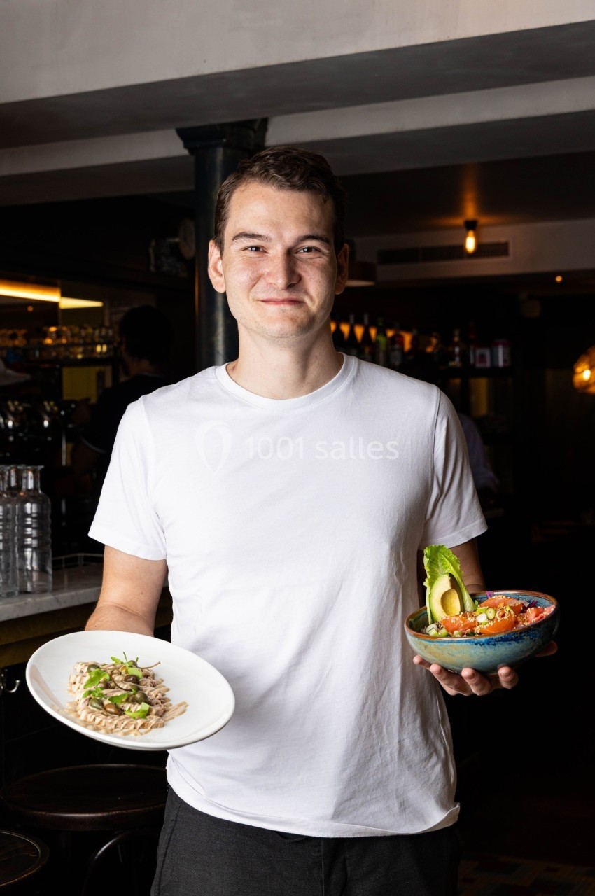 Un homme souriant tenant un plat de risotto et un bol de salade garnie d'avocat dans un restaurant.