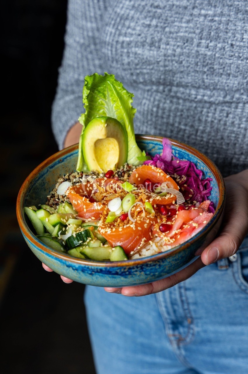 Bol coloré contenant du saumon, avocat, légumes frais, quinoa et graines, tenu par une personne en jean et pull gris.
