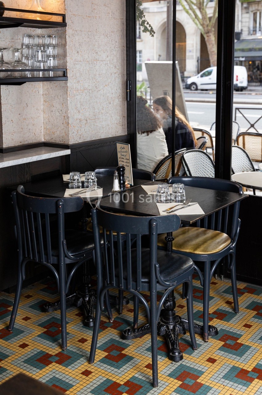 Table de café avec chaises noires, verres et menu, située près d'une grande fenêtre donnant sur une rue animée.
