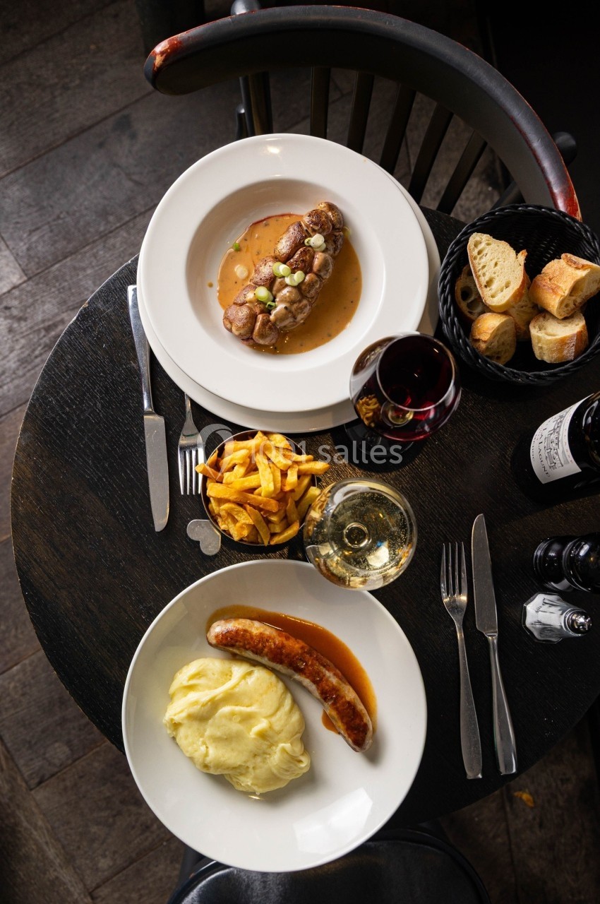 Repas composé de saucisse-purée et entrecôte avec frites, accompagné de pain, vin rouge et blanc sur une table en bois.