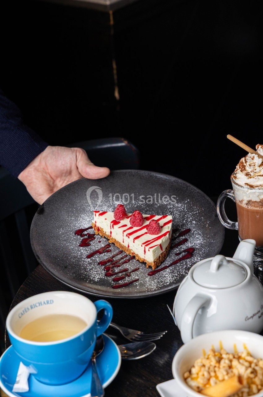 Une main sert une assiette avec une part de cheesecake décorée de framboises et coulis, accompagnée de boissons chaudes.