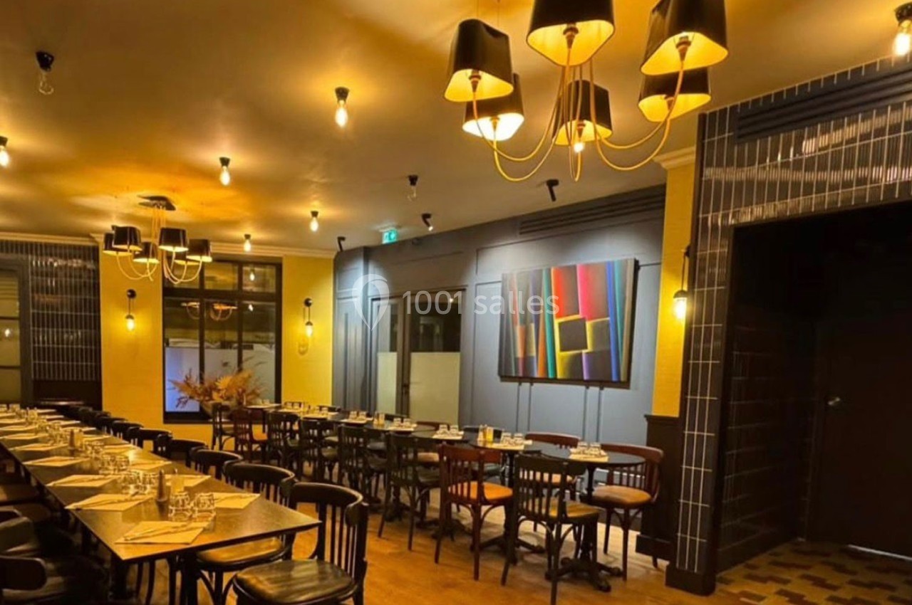 Salle de restaurant chaleureuse avec tables alignées, chaises en bois, éclairage tamisé et tableau coloré au mur.