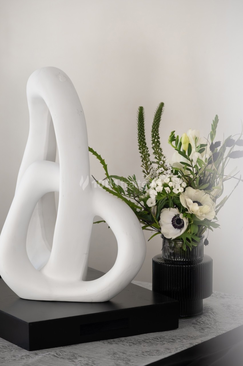 Sculpture blanche abstraite sur socle noir à côté d'un vase noir contenant un bouquet de fleurs blanches et vertes.