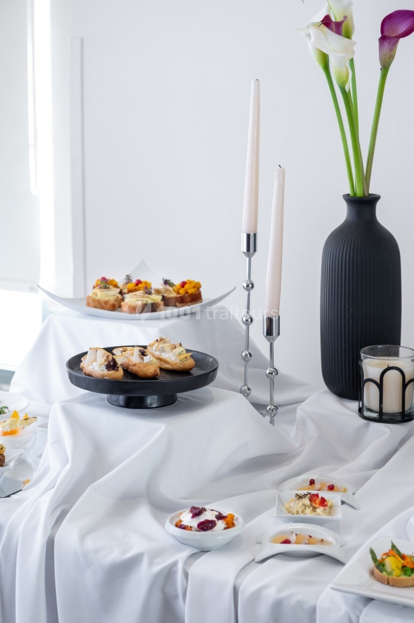 Table dressée avec des amuse-bouches variés, bougies allumées et vase noir contenant des fleurs blanches et violettes.