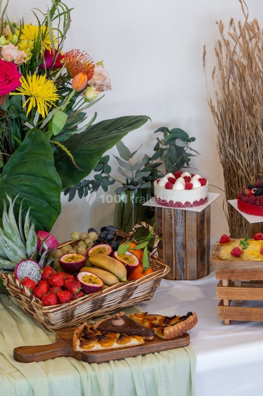 Plateau de fruits exotiques, tarte aux fruits et gâteau décoré de framboises, disposés sur une table décorée de fleurs.